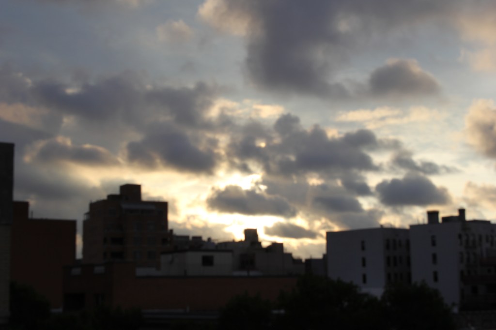 a blurry summer sunrise, yellow with gray clouds billowing in front of it, in front of the silhouetted rooftops of  astoria, looking east
