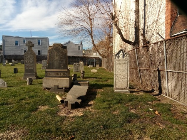 gravestones in a small yard next to a building with two story homes in the background. a gnarly tree stretches across the middle of the frame